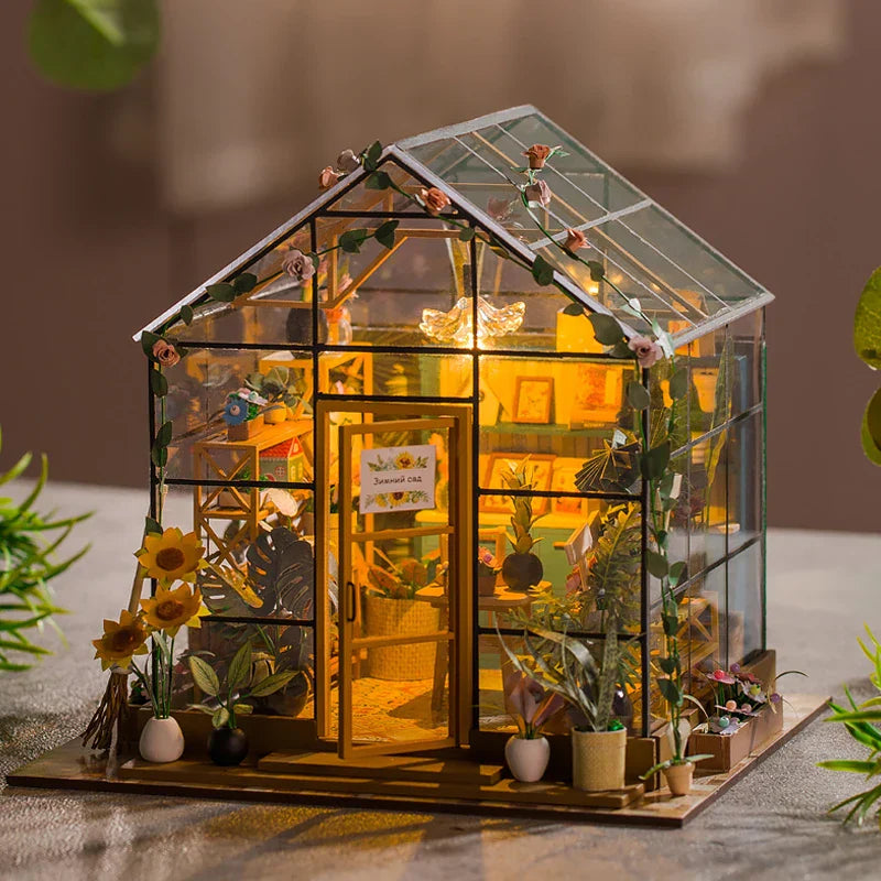 A miniature greenhouse with transparent walls and a gabled roof, illuminated from the inside, surrounded by various miniature plants and figures.