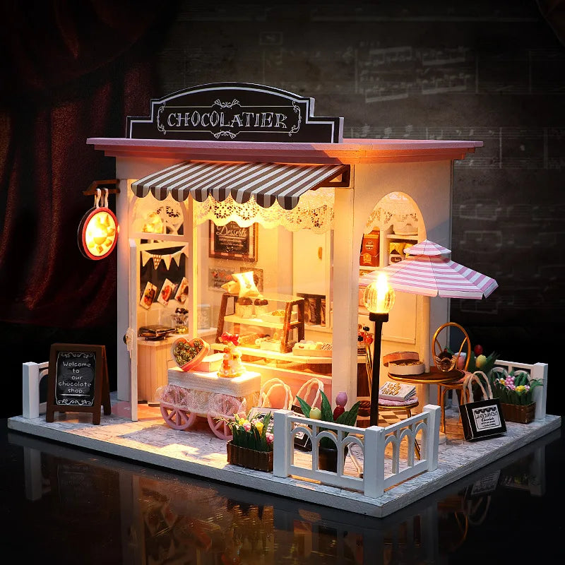 A miniature greenhouse with transparent walls and a gabled roof, illuminated from the inside, surrounded by various miniature plants and figures.