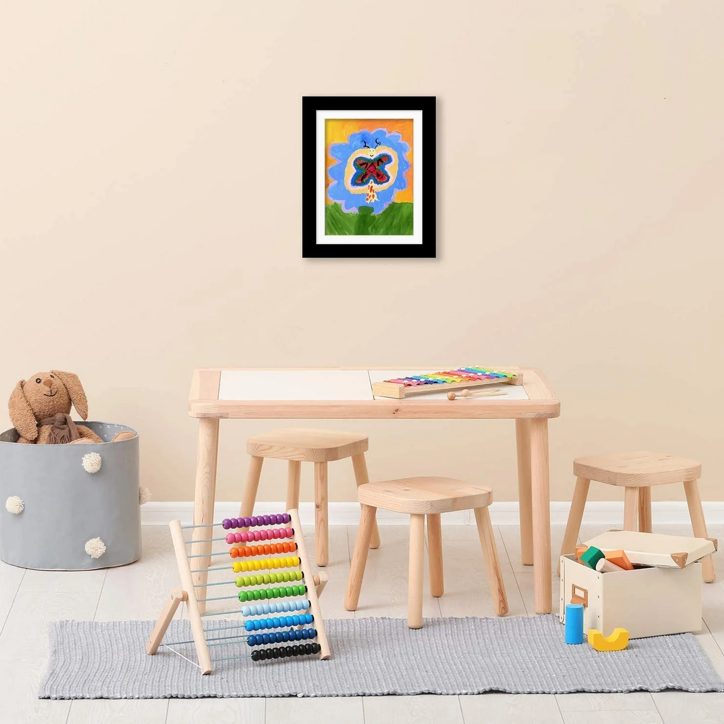 Children's play area with a table, stools, and toys against a beige wall with a framed picture.