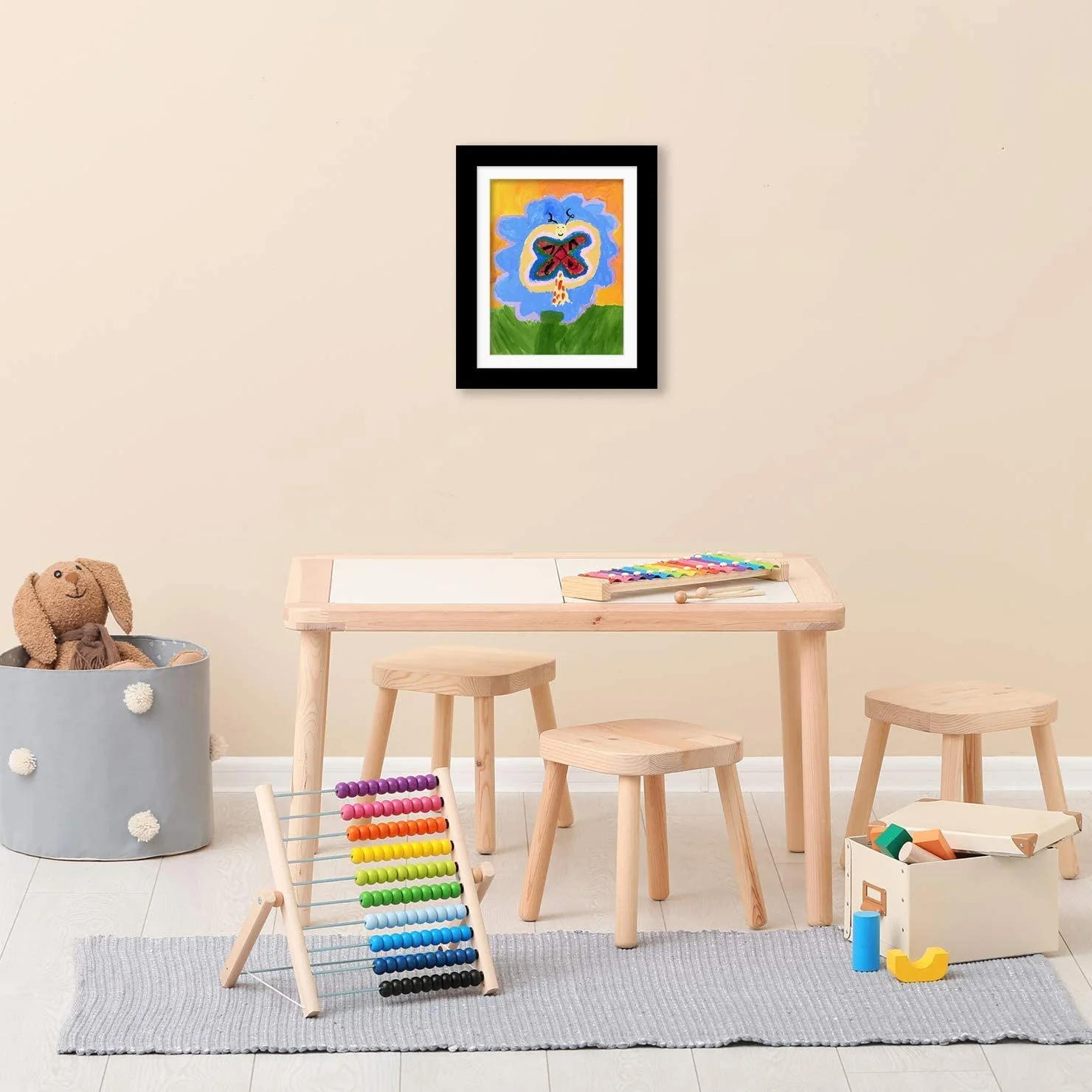 Children's play area with a table, stools, and toys against a beige wall with a framed picture.